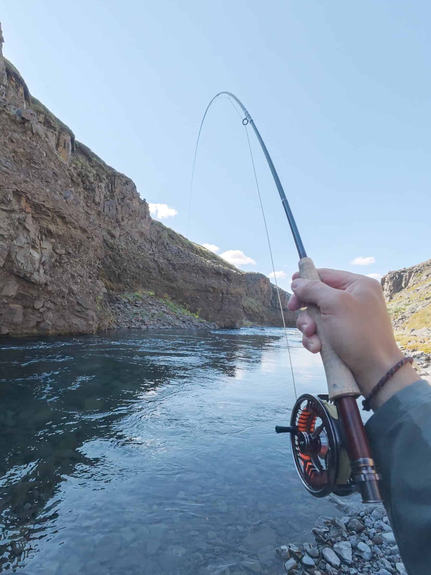 Person fishing in Icelandic river with a spinning rod and scenic cliffs.