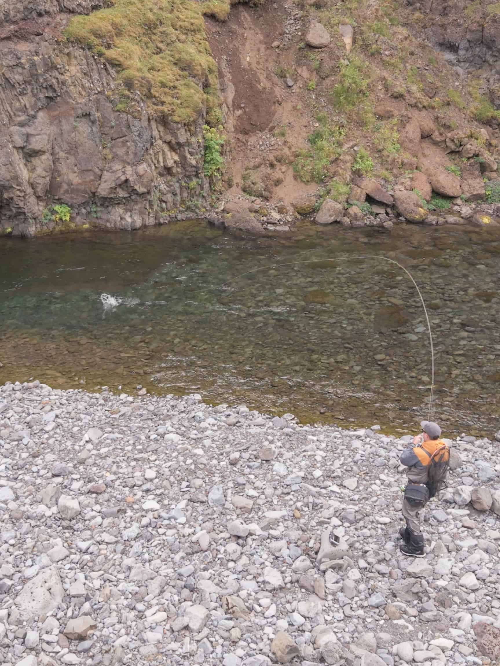 Angler fishing in Iceland's river with rocky shore and steep cliffs.