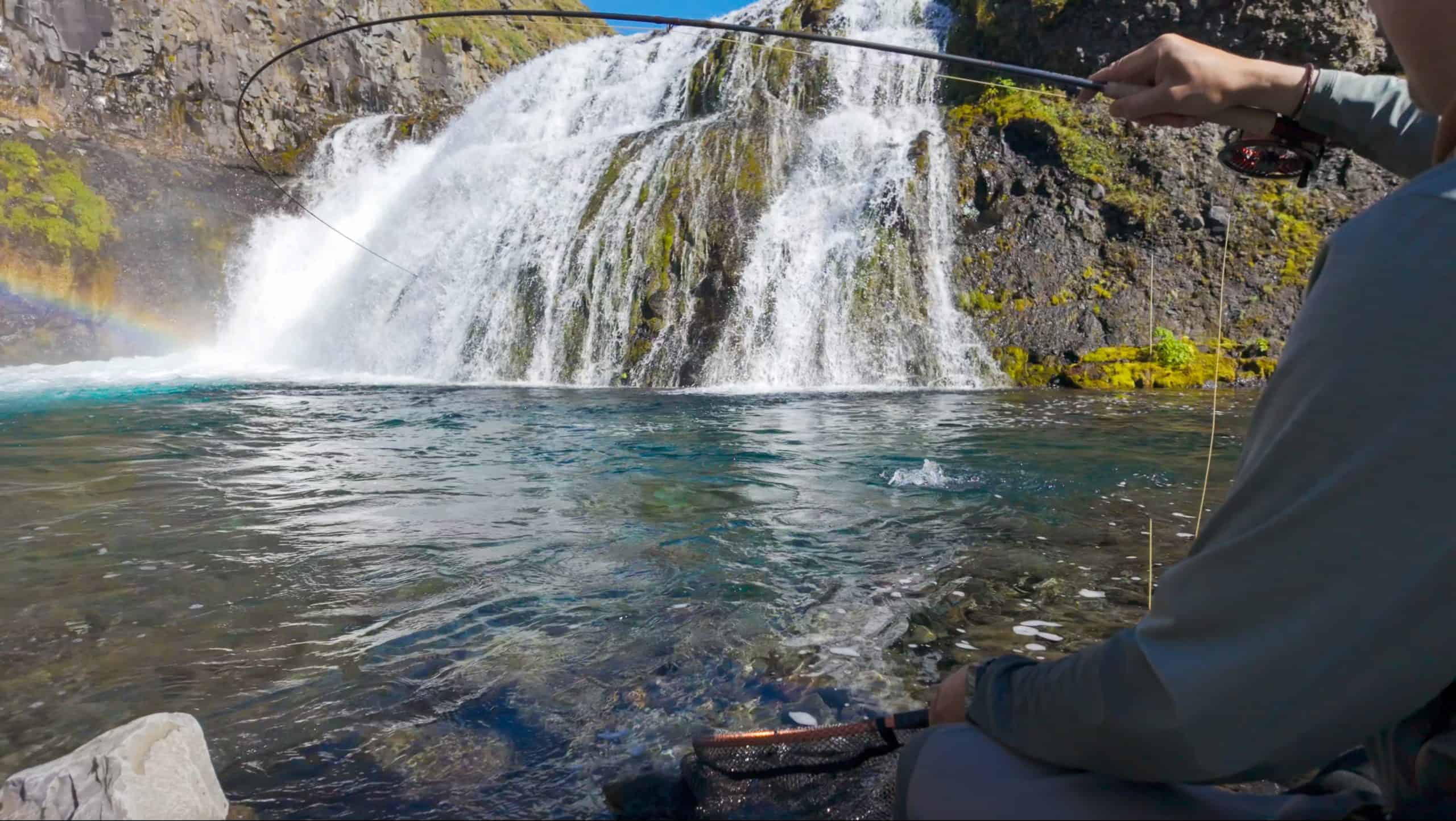 Angler fishing near a waterfall in Iceland with clear water and lush surroundings.