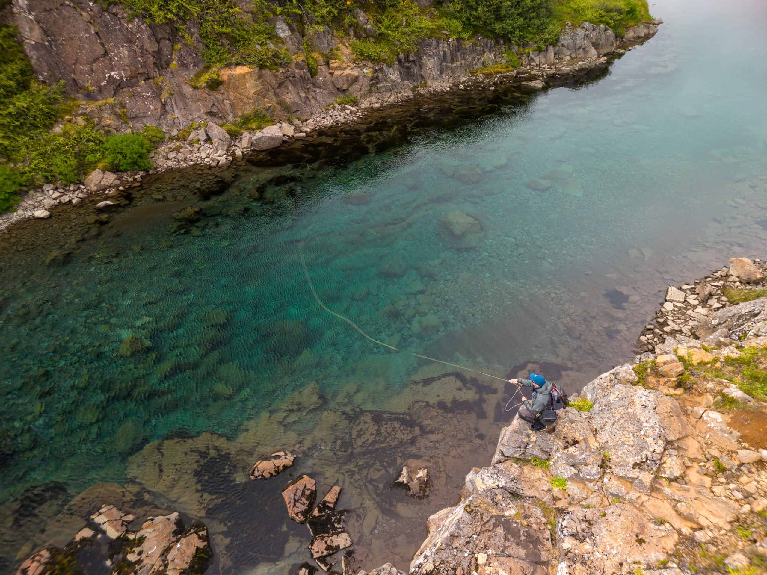 Fisherman fishing in clear river with rocky banks and lush greenery.