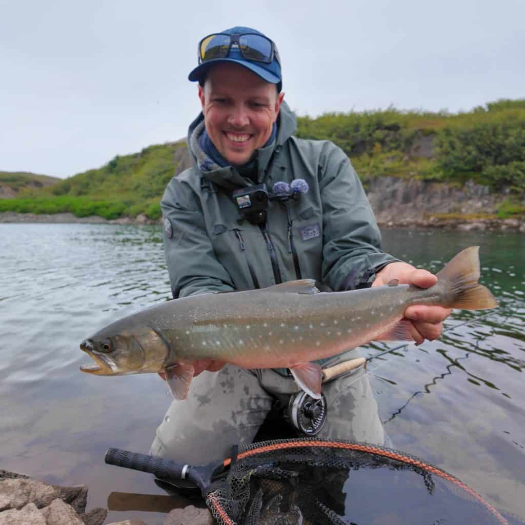 Smiling angler holding a large Arctic Char by the riverbank.