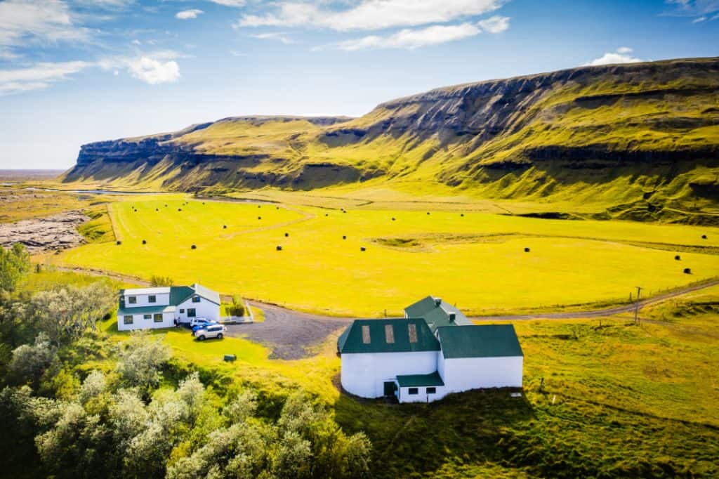 Icelandic farmhouses at the base of a green, layered mountain under a blue sky.
