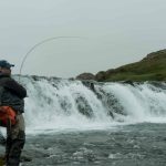 A fisherman standing near a rushing waterfall fishing in a natural river landscape. Fishing enthusiast casting line in a river waterfall, enjoying outdoor sport and fresh water fishing.