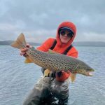 Angler holding a large rainbow trout during fishing trip, dressed in outdoor gear with cloudy sky background. Big rainbow trout caught by angler in outdoor water setting.