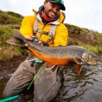 A smiling angler in outdoor gear displaying a freshly caught colorful trout in a freshwater river. Spinning fisherman holding large wild trout in a river, showcasing fishing success and outdoor adventure.