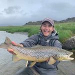 Joyful fisherman showcasing a big catch of trout in natural outdoor setting. Vibrant young angler holding a large spotted fish near riverbank.