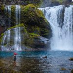 Scenic waterfall with fishing activity in pristine Icelandic nature, highlighting freshwater fishing opportunities and outdoor adventure. Vibrant waterfall scene with a person fishing in clear river waters, surrounded by moss-covered rocks and lush natural landscape.