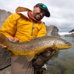 Happy angler holding a large brown trout near a river with rocky cliffs under a cloudy sky. Brown trout caught by an angler in a river with rocky surroundings on a cloudy day.
