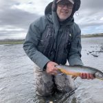 A smiling man holding a fish he caught while fly fishing in a river, dressed with outdoor gear and weatherproof clothing. Freshly caught fish angler in outdoor river fishing trip, showcasing fishing gear and experience.