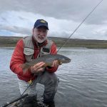 A smiling fisherman proudly displays his catch of a rainbow trout in a scenic river setting. Rainbow trout fishing outdoors in a serene river landscape.