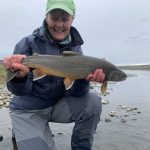 Smiling angler holding big fish by river, enjoying fishing outdoors with scenic landscape. Freshwater catch, man holding a large fish by the river, fishing trip, angler with fishing gear, outdoor recreational activity.