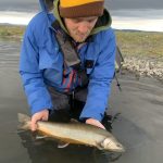 Man in blue jacket holding a fish while fishing in a river during overcast weather. Vibrant image of man in outdoor fishing gear holding a freshly caught fish in a river setting.