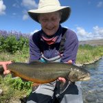 Beautiful outdoor scene of a woman holding a rainbow trout by a river with wildflowers and blue sky, emphasizing fishing and nature. Rainbow trout caught during a fishing trip in a scenic river setting, showcasing recreational fishing and outdoor adventure.