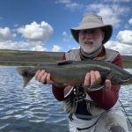 A smiling angler proudly displays a big fish caught while fishing in a tranquil lake under a blue sky with clouds. Caught large fish during outdoor fishing adventure in scenic lake environment.