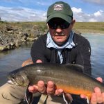 Man showing off freshly caught rainbow trout during outdoor fishing trip, river background. Viral Fish Partner logo on cap, man holding a large rainbow trout by river, outdoor fishing scene.