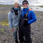 Two smiling anglers in waterproof fishing gear standing on rocky riverbank with a scenic view of a river and mountains in Iceland. Fishing partners enjoying outdoor fishing adventure in Iceland, wearing outdoor gear and waders by riverbank.