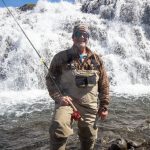 Happy angler enjoying fishing adventure near a waterfall with fishing rod and gear. Fishing enthusiast by waterfall with fishing gear and outdoor clothing.