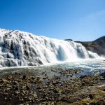 Beautiful cascading waterfall in Iceland's wilderness, with rocky foreground and bright sunlight. Vibrant waterfall cascading over rocks under clear blue sky, showcasing natural scenic beauty.