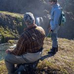 People fishing at a picturesque waterfall with rugged terrain and vibrant nature, highlighting outdoor fishing adventures. Fishing enthusiasts enjoying a scenic river fishing trip with waterfall views in the background, dressed in outdoor gear.
