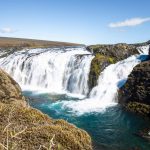 Scenic Icelandic waterfall in daylight showcasing powerful cascading water and natural landscape. Vibrant waterfall flowing over rugged rocks with lush green surroundings and bright blue sky.