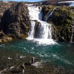 Breathtaking Icelandic waterfall scene with anglers fishing in a river at the base of a stunning waterfall. Flowing waterfall surrounded by rocky landscape and two anglers fishing in Iceland.