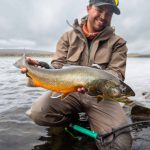 Happy man holding large fish caught from river, outdoors fishing activity, and rural landscape background. Vibrant wild fish caught during fishing trip, showcasing angler's success in freshwater fishing.