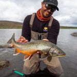 Smiling angler with sunglasses holding a large wild brown trout in a scenic river setting for fishing adventures. Caught fish in freshwater river, angler holding colorful trout, outdoor fishing adventure, fish species, fishing gear, nature, success, excitement, Fish Partner.