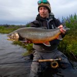 Beautiful catch of a freshwater fish near the riverbank on a cloudy day. Vivid image of a man holding a large fish while fishing in a river with lush greenery and flowers in the background.