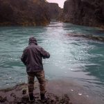 Angler standing on rocks in canyon river, fishing in clear blue water surrounded by rugged cliffs. Fishing in a scenic river canyon, casting a line in pristine waters for freshwater fish.