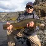 Smiling angler holding a freshly caught rainbow trout by a riverbank, outdoor fishing trip. Rainbow trout fishing in Iceland, catch and release sport fishing adventure.