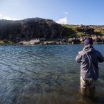 Engaged in freshwater fishing at a picturesque river surrounded by rugged terrain and nature. Fishing enthusiast casting a line in a scenic river with rocky cliffs and blue sky.