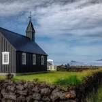 Charming black church situated in Icelandic countryside with scenic views of Mount Etna and lush green fields. Old black church in a rural landscape with Mount Etna in the background, Icelandic scenery, and green fields, under dramatic cloudy skies.