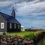 Charming black church situated in Icelandic countryside with scenic views of Mount Etna and lush green fields. Old black church in a rural landscape with Mount Etna in the background, Icelandic scenery, and green fields, under dramatic cloudy skies.