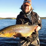 A content angler displays a massive rainbow trout caught in scenic waters, demonstrating successful freshwater fishing. Large rainbow trout fishing, caught in pristine waters, showcasing expert angling techniques for freshwater fish.