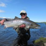 A fisherman proudly displays a large salmon he caught in a pristine river with mountainous landscape. Large Atlantic salmon caught by a fisherman in a scenic river with snow-capped mountains in the background.