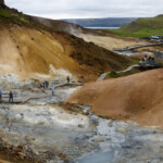 Steam and mineral-rich waters flow through the geothermal landscape, attracting visitors exploring the natural wonder at the geothermal park. Volcanic geothermal area with steaming vents, colorful mineral deposits, and visitors walking along wooden pathways.