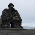 Rustic stone sculpture of a bearded figure overlooking the sea, crafted from layered rocks for outdoor art display. Weathered stone sculpture of a bearded figure on a coastal site, made from stacked stones.