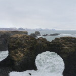 Majestic Icelandic coastal rock formation with arches and rugged black volcanic terrain along the Atlantic Ocean. Dramatic seascape and mountain backdrop. Bold mountain coastal arch with rugged volcanic rocks and wild ocean waves in Iceland. Scenic natural landscape with snowy mountains in background.