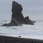 A dramatic ocean scene featuring large sea stacks, rocky shoreline, and turbulent waters. Rugged coastal rocks with crashing waves under an overcast sky.