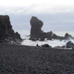 Beautiful volcanic landscape with ocean waves crashing against jagged rocks and distant sea stacks. Dark rocky coastline with sea stacks and black pebble beach near ocean waves.