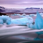 Colorful ice formations create a stunning Arctic or Antarctic landscape scene. Icebergs floating in a cold, glacial lagoon with snow-covered mountains in the background.