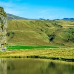 Foggy river valley with cliffs and waterfall in the distance, lush green landscape and clear blue sky, rural Icelandic scenery.