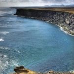 Scenic coastal cliffs with ocean waves hitting the shoreline. Rich coastal cliffside overlooking a deep blue ocean cove.