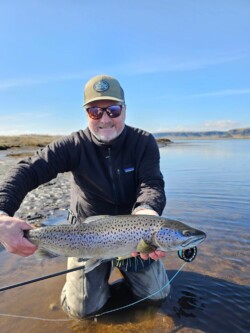 Happy man in outdoor fishing gear holding a big rainbow trout by a calm river with mountains in the background. Large rainbow trout caught in a scenic river during fishing trip, showcasing successful freshwater angling.