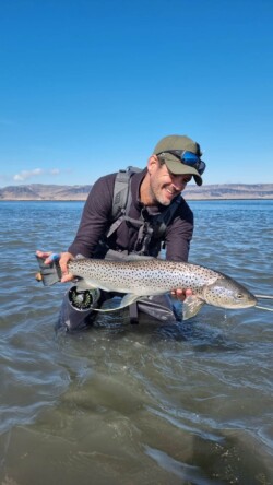 A smiling man holds a large rainbow trout he caught while fishing on a lake with scenic mountains in the background. Vibrant rainbow trout catch in freshwater fishing lake.