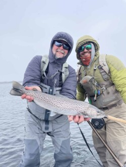 Two fishermen in outdoor gear proudly display a rainbow trout caught during a fishing trip. Vancouver rainbow trout fishing with anglers holding a freshly caught fish in a scenic water setting.