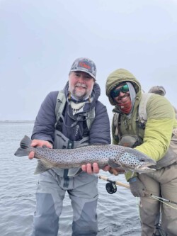 Smiling anglers showcase a large, freshly caught rainbow trout on a fishing trip setback on a lake or river. Vibrant trout caught during fishing trip with two anglers in a cold, overcast environment.