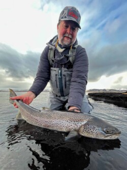 Man with fishing gear holding a large rainbow trout fish in a river. Brighton man holding a large fish in freshwater river during overcast weather.