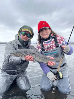 Anglers showcasing a successful trout catch during a fishing trip in cold weather. FSH-001: Two anglers in waterproof gear holding a freshly caught rainbow trout by a river in overcast weather.