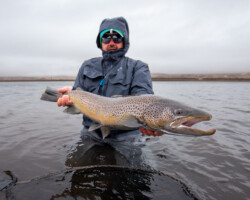 A fisherman proudly displaying a large brown trout in a river with overcast skies. Large brown trout caught by angler in outdoor river setting, fishing tour, sport fishing, fishing equipment, overcast sky.